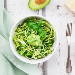 Zoodles con pesto di avocado, guarniti con pinoli e basilico. Accanto, mezzo avocado, un pezzo di parmigiano e una forchetta.