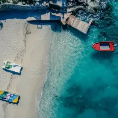 Widok z lotu ptaka na plażę w Albanii. Kilka łodzi, w tym czerwona, zacumowana przy drewnianym pomoście, czeka na turystów.