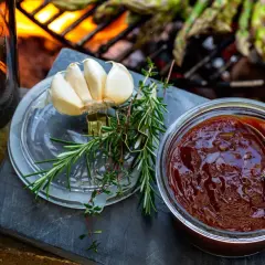 En burk med hemlagad bbq sås, vitlök och färska örter förbereds för grillen där sparris redan ligger på gallret.