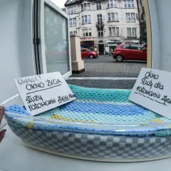 Okno życia z tabliczkami informacyjnymi w koszu, widok na ulicę z budynkami i samochodami.