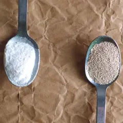 Deux cuillères sur fond de papier kraft : l'une avec de la poudre blanche (levure chimique), l'autre avec des granulés beiges (levure boulangère).