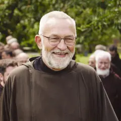 Bruder Paulus, ein älterer Mann mit Brille und Bart, lächelt in die Kamera. Hinter ihm stehen weitere Männer in braunen Kutten.