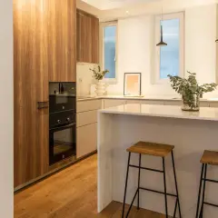 Cocina moderna con muebles de madera, encimera blanca y dos taburetes. La luz fría k ilumina el espacio, creando un ambiente acogedor.