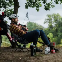 Mężczyzna odpoczywa podczas wycieczki rowerowej, gotując posiłek na kuchence turystycznej. Obok rower z sakwami.