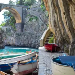 Boats line the shore of Fiordo di Furore, a stunning fjord with turquoise waters and a dramatic arched bridge overhead.