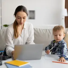 Kobieta pracuje na laptopie, podczas gdy jej syn rysuje obok. To codzienna opieka na dziecko i praca zdalna w jednym.