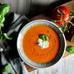 En skål med krämig tomatsoppa, gjord på krossade tomater, toppad med en klick gräddfil och basilika. En färsk tomat och en flaska Knorr Fresh Vegetable Vegan Fond ligger bredvid.