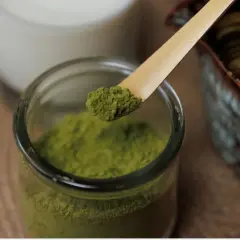 Sypka zielona matcha na bambusowej łyżeczce, obok słoiczek z proszkiem, szklanka mleka i miotełka do ubijania. Idealne składniki, by dowiedzieć się, jak pić matcha.