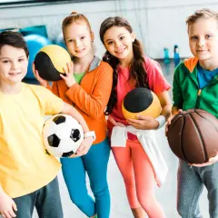 Cztery uśmiechnięte dzieci w sportowych ubraniach trzymają piłki: nożną, do koszykówki i dwie treningowe.