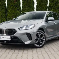 Szare BMW 118d z czarnym grillem i felgami stoi przed żywopłotem.
