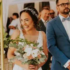 Szczęśliwa panna młoda z bukietem kwiatów podczas szybkiego ślubu cywilnego. Pan młody w niebieskiej marynarce czeka na przysięgę.