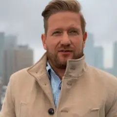 Bartek Jędrzejak w płaszczu na tle miasta. Marzy o powrocie do żony i dzieci po pracy.