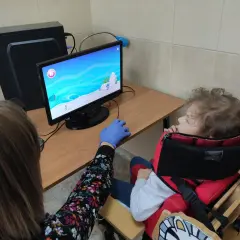 Dziecko w specjalnym foteliku siedzi przed komputerem, na ekranie gra. Obok siedzi osoba w rękawiczce.