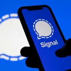 Dłoń trzyma smartfon z otwartą aplikacją Signal. W tle widoczne logo Signal.