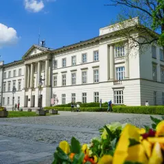 Biały budynek z kolumnami i zielenią, w tle błękitne niebo z chmurami.
