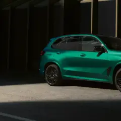 Zielony BMW X5 M z czarnymi felgami stoi na tle ciemnej architektury, oświetlony pasem światła.