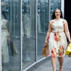 Uśmiechnięta kobieta w kwiecistej sukience plus size, z torbami zakupowymi w rękach, idzie obok witryny sklepowej.