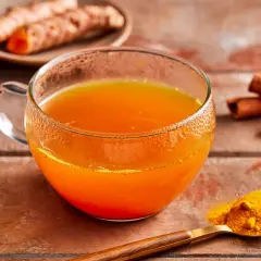 Ein Glas leuchtend orangefarbener Kurkuma-Tee steht auf einem Holztisch. Daneben liegen frische Kurkumawurzeln, Zimtstangen und ein Löffel mit Kurkumapulver. Die Wirkung von Kurkuma-Tee ist bekannt.
