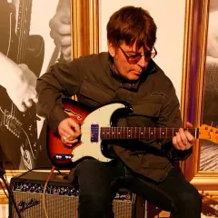 Mężczyzna w okularach gra na gitarze elektrycznej Fender, siedząc na wzmacniaczu.