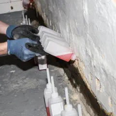 Hände mit Handschuhen führen kleine Flaschen mit roter Flüssigkeit an eine Wand, um einen Lichtschacht abzudichten.
