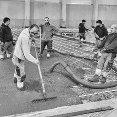 Arbeiter verteilen frisch gegossenen stahlfaserbeton bodenplatte mit Rechen. Beton wird aus Schlauch gepumpt.