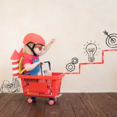 Dziecko w wieku przedszkolnym w koszyku na kółkach, z rakietą na plecach, symbolizuje rozwój fizyczny i wyobraźnię.