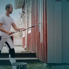 Man målar om huset med en lång roller. Det röda trähuset får nya gråa paneler.