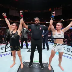 Two female fighters stand with arms raised, a referee between them. The outcome of their boxing draw is announced to the roaring crowd.