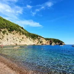 Kamienista plaża w Czarnogórze z turkusową wodą i zielonym wzgórzem w tle.