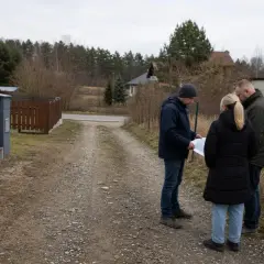 Trzy osoby omawiają mapę na żwirowej drodze. Widać domy i ogrodzenia, a także oceniany jest odległość domu od drogi służebnej.