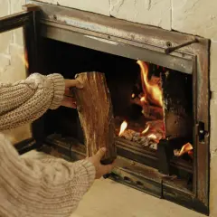 Une personne en pull ajoute une bûCHE dans un poêle à bois qui fume, porte ouverte. La chaleur des flammes réchauffe la pièce.
