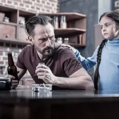 Smutna dziewczynka kładzie rękę na ramieniu ojca, który pije alkohol. To obraz trudnej sytuacji rodzinnej, która może wymagać terapii DDA.