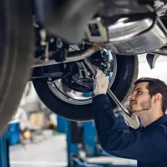 Meccanico con guanti bianchi lavora sotto un'auto sollevata, cercando la causa di un rumore metallico da sotto la macchina.