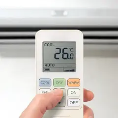 Dłoń naciska przycisk na pilocie klimatyzacji, wybierając funkcje chłodzenia. Na ekranie widoczna temperatura 26°C.
