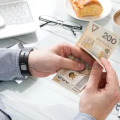 Ręce liczą polskie banknoty 200 zł. Obok laptop, kawa, rogalik i notatnik. To obrazek pokazujący klasa średnia zarobki.