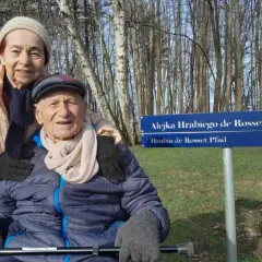 Uśmiechnięta para seniorów pozuje przy tablicy "Alejka Hrabiego de Rosset" w dom seniora Rotmanka.