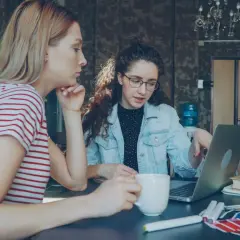 Dwie kobiety pracują nad projektem w biurze. Jedna z nich, z burzą kręconych włosów, wskazuje coś na laptopie. To może być początek ich wspólnej spółki jawnej.