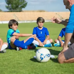 Trener piłki nożnej instruuje młodych zawodników siedzących na murawie.