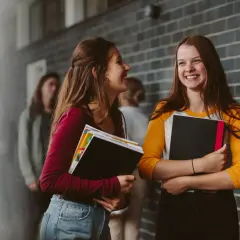 Dwie uśmiechnięte uczennice trzymające książki na tle ceglanej ściany.