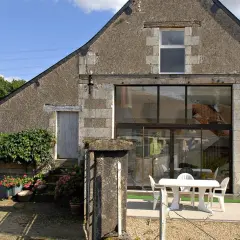 Ancienne grange en cours de rénovation, avec une façade en pierre et un grand mur vitré moderne. Une table et des chaises blanches sont installées sur la terrasse.