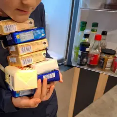 Dziecko trzyma stos opakowań masła przed otwartą lodówką z butelkami sosów.