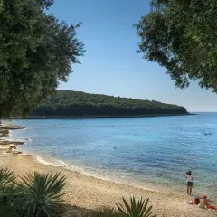 Słoneczny dzień na plaży w Porto Sole. Ludzie odpoczywają na kamienistym brzegu, a w krystalicznie czystej wodzie widać kąpiących się.