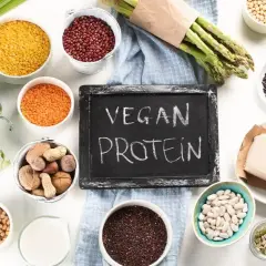 Tablica z napisem "VEGAN PROTEIN" otoczona różnorodnymi roślinnymi źródłami białka: soczewicą, fasolą, brokułem, szparagami, tofu i orzechami.