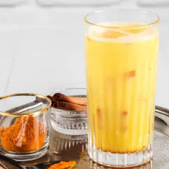 Ein Glas goldene Milch mit Kurkumapulver und Zimtstangen daneben.
