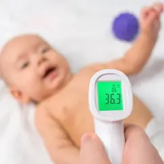 Dziecko leży na białym tle, a ręka trzyma termometr bezdotykowy. Na ekranie termometru widoczna jest temperatura 36.3°C, co oznacza, że temperatura mierzona w odbycie jest prawidłowa.