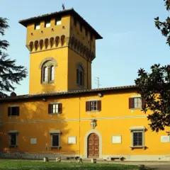Edificio giallo con torre merlata a Borgo San Lorenzo, circondato da alberi verdi e un cielo azzurro.