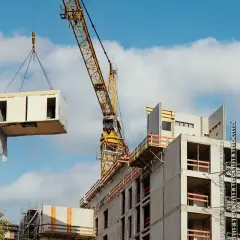 Żółty dźwig budowlany podnosi modułowy element drewniany na placu budowy. Betonowy budynek wznosi się w tle, a niebo jest błękitne z chmurami.
