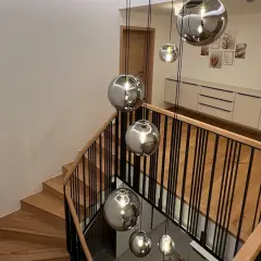 Nowoczesne oświetlenie klatki schodowej w bloku: wiszące, lustrzane kule tworzą efektowną instalację nad drewnianymi schodami z czarną balustradą.