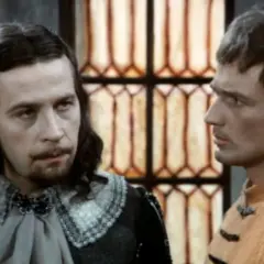 Dwóch mężczyzn w historycznych strojach, jeden z długimi włosami i brodą, drugi w pomarańczowej tunice. W tle okno z witrażem, jakby czarne chmury obsada.