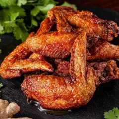 A pile of golden-brown, crispy alette di pollo, glistening with sauce, are served on a dark slate. Coriander, ginger, garlic, and peppercorns complete the appetizing presentation.